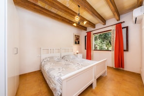 A bedroom with a wooden ceiling, a bed with a patterned bedspread, a bedside table with a lamp, and a window with red curtains.