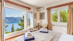 Une chambre d’hôtel comprenant un lit, deux chaises, une table et de grandes fenêtres offrant une vue sur la mer et les montagnes.