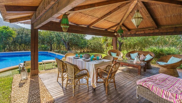 Un espacio al aire libre techado con piscina, mobiliario de mimbre y una mesa preparada para comer.