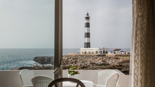 Vista de un faro desde una habitación con sillas blancas y una mesa, con vistas al mar y al paisaje rocoso.
