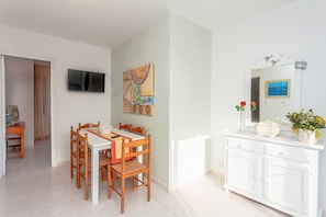 Un comedor con una mesa y sillas, un televisor montado en la pared y un armario blanco con un espejo y objetos decorativos.