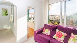 A purple sofa with floral cushions, a white wall, and a balcony with a view of buildings and greenery. - Apartment Cala en Forcat 36 with Balcony & Shared Pool (Cala en Blanes)