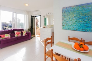 Una sala de estar con un sofá morado, una mesa de comedor con un plato de naranjas y un ventanal con vistas a zonas verdes.