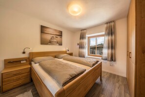 A bedroom with two wooden beds, a nightstand, a wall-mounted lamp, a painting, and a window with curtains.