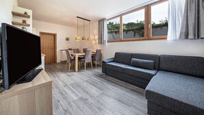 Ein modernes Wohnzimmer mit Flachbildfernseher, grauem Sofa, Esstisch mit Stühlen und einem großen Fenster mit Vorhängen.