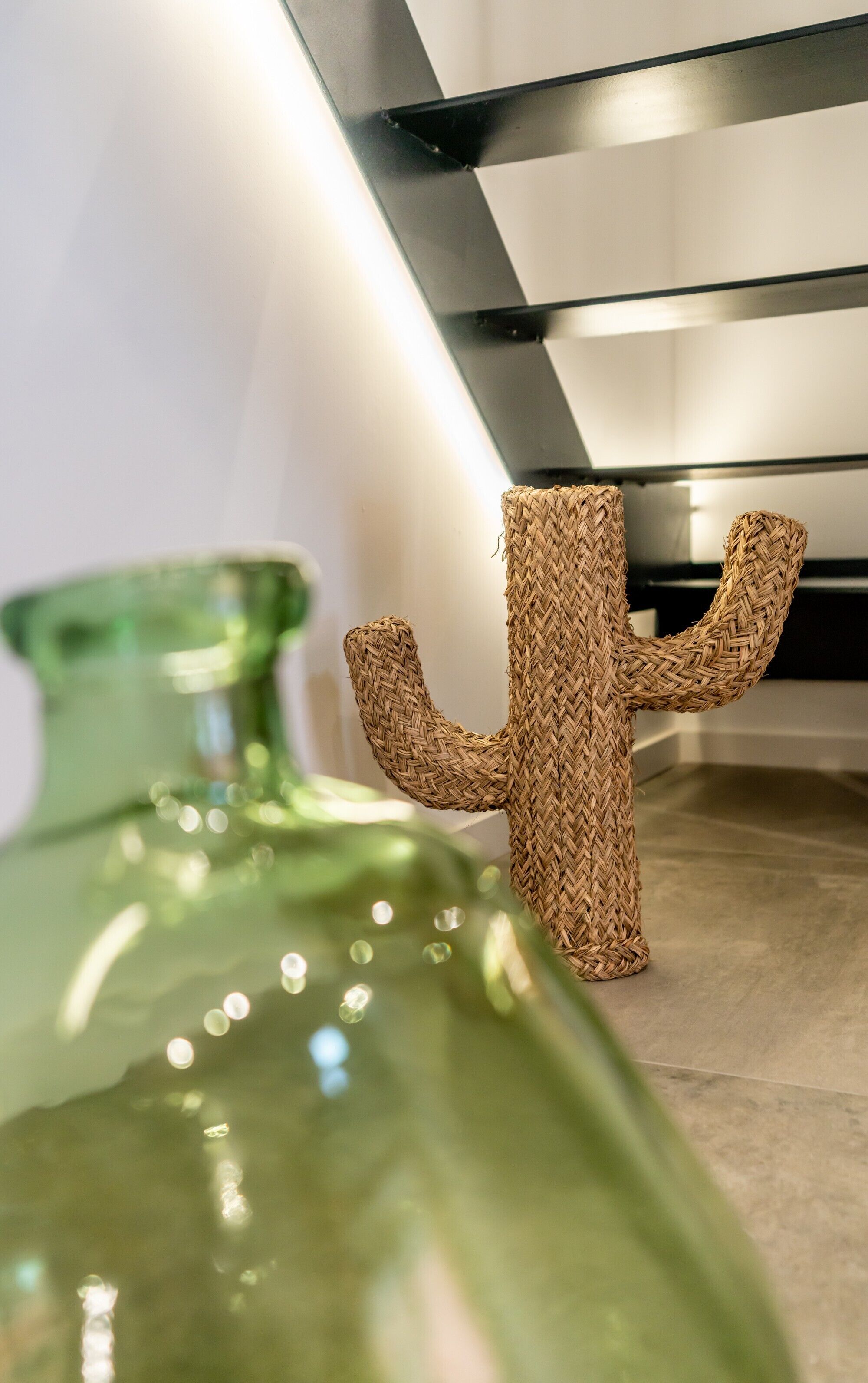 Un vase en verre vert et une sculpture de cactus en fibres tressées, posés sur un sol carrelé.