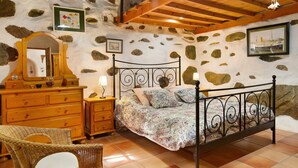 Ein Schlafzimmer mit Steinwand, Holzbalkendecke, einem Bett mit Metallgestell, einem Holzkleiderschrank, einem Korbsessel und einem gerahmten Bild an der Wand.
