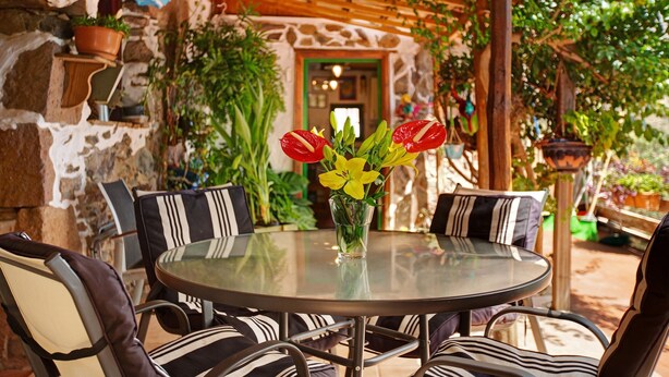 Ein runder Glastisch mit gestreiften Stühlen, eine Vase mit gelben und roten Blumen und eine Steinwand mit einer hölzernen Pergola darüber.