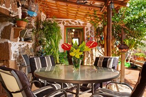 Ein runder Glastisch mit gestreiften Stühlen, eine Vase mit gelben und roten Blumen und eine Steinwand mit einer hölzernen Pergola darüber.
