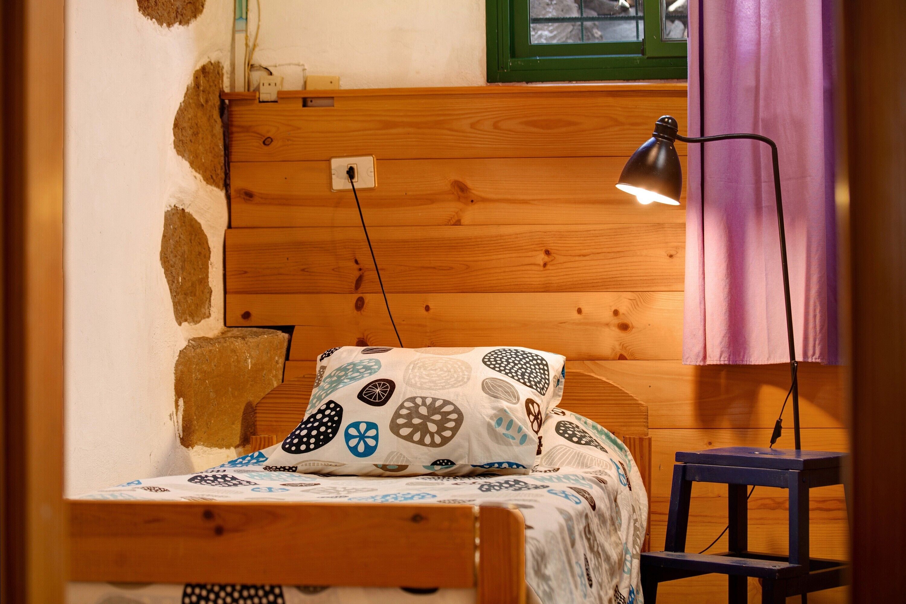 A wooden bed with a patterned pillow, a bedside lamp, and a small wooden stool.