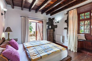 A bedroom with a bed, wooden beams, a window with curtains, a lamp, and a stone wall.