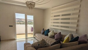 A living room with a beige sofa, patterned cushions, a chandelier, and a sliding glass door leading to a balcony. - 2 Bed Flat - Sleeps 5 - Pets - Pool - Parking (Saidia)