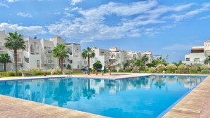 A swimming pool with a tiled deck, surrounded by palm trees and white buildings with red domes. - 2 Bed Flat - Sleeps 5 - Pets - Pool - Parking (Saidia)