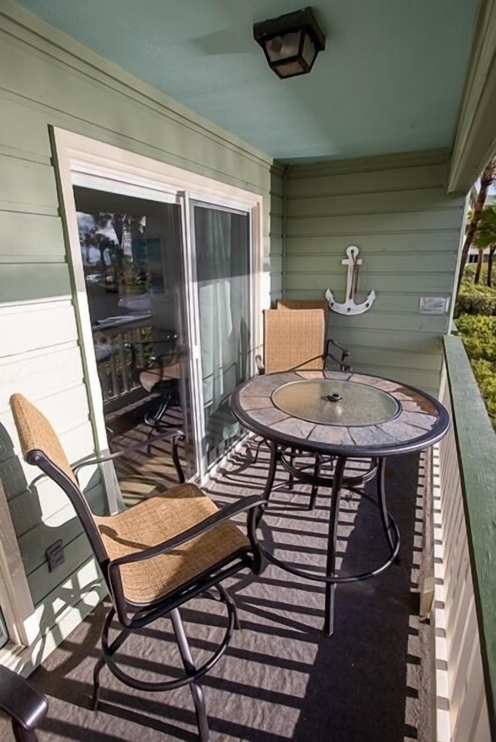 Een overdekt terras met een ronde tafel en stoelen, een schuifdeur en een kleine ankerdecoratie aan de muur.