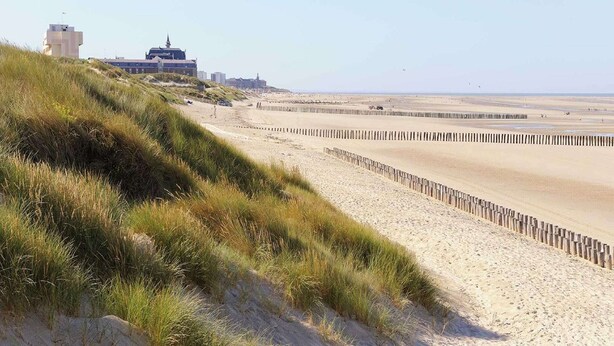 Une plage avec un sentier sablonneux et des dunes herbeuses, une clôture en bois et des bâtiments en arrière-plan.