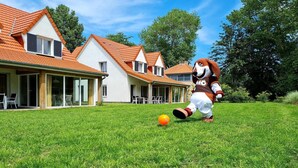 Une mascotte se trouve dans un espace verdoyant, avec une maison en arrière-plan.
