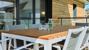 Une table en bois placée à l’extérieur, avec des chaises blanches, un vase en verre contenant une plante et des verres transparents.