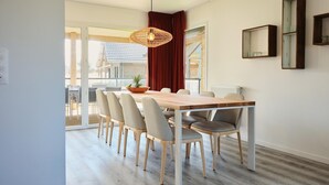 Un espace repas moderne avec une table en bois et six chaises, un luminaire suspendu et une vue sur l’extérieur grâce à de grandes fenêtres.