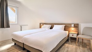 Un lit double avec une tête de lit en bois, des tables de chevet et une chaise, dans une pièce bien éclairée dotée d’une fenêtre et de rideaux.