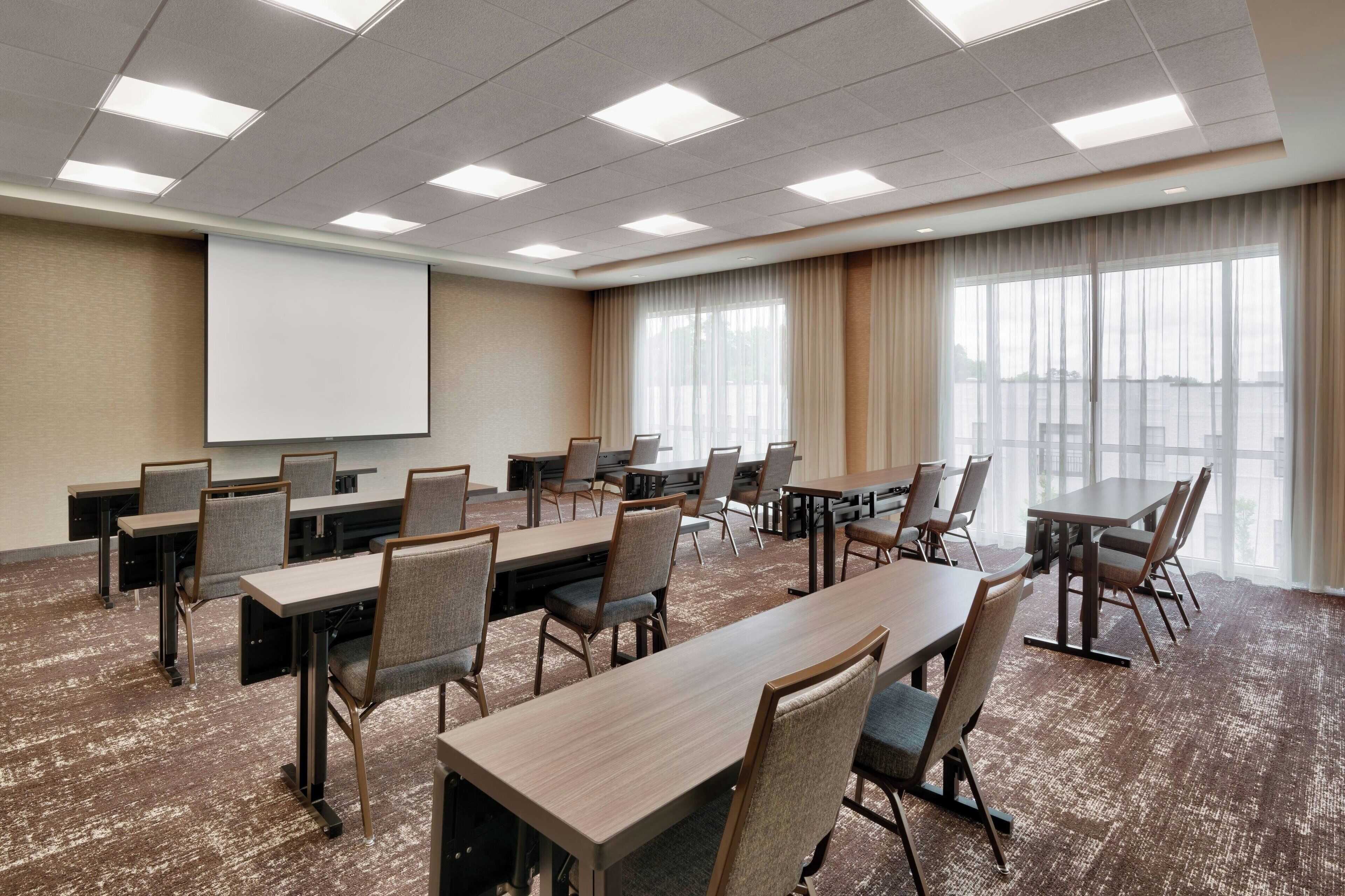 A conference room with multiple tables and chairs, a projector screen, and large windows with curtains.