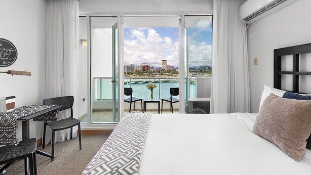 Une chambre d’hôtel avec un lit, un bureau avec deux chaises, et une vue sur le paysage urbain et l’eau, grâce à de grandes portes vitrées.