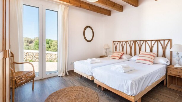 A bedroom with two beds, a wicker chair, a round rug, and a view through a glass door. - Finca Na Bona · Finca Na Bona. Perfect location. Menorca (Mahón)