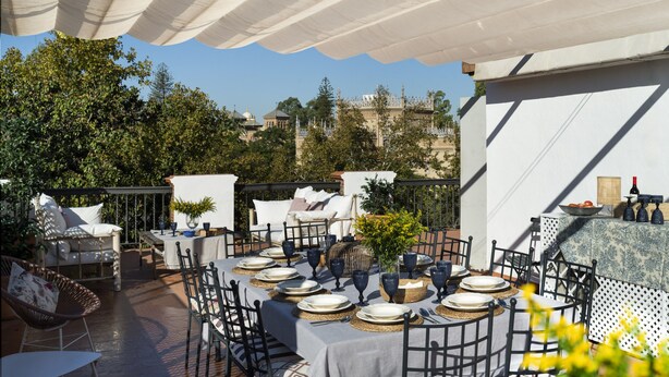 An outdoor dining area with tables set for four, surrounded by chairs and umbrellas, overlooking a scenic view of a historic building. - Villa Alfonso, Luxurious restored palace house next to Plaza de España. (Sevilla)
