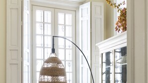 A dining area with a round table set for breakfast, wicker chairs, and a large window with white shutters. - Villa Alfonso, Luxurious restored palace house next to Plaza de España. (Sevilla)