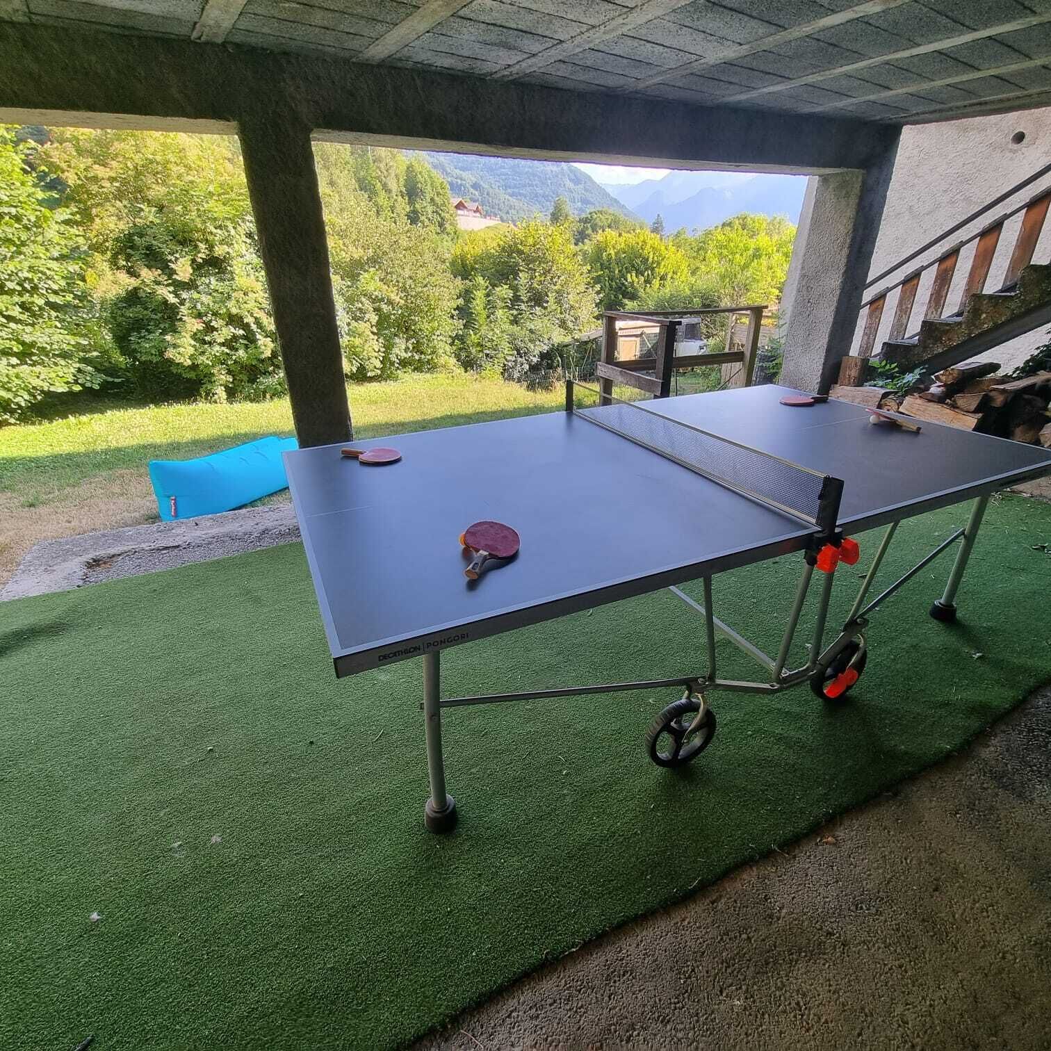 Ett bordtennisbord med två racketar och en boll, placerat på konstgräs under en betongkonstruktion med utsikt över ett bergigt landskap.
