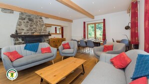 Ein gemütliches Wohnzimmer mit Kamin aus Stein, grauen Sofas mit roten Kissen, einem Holztisch und einem Essbereich mit Blick ins Grüne.