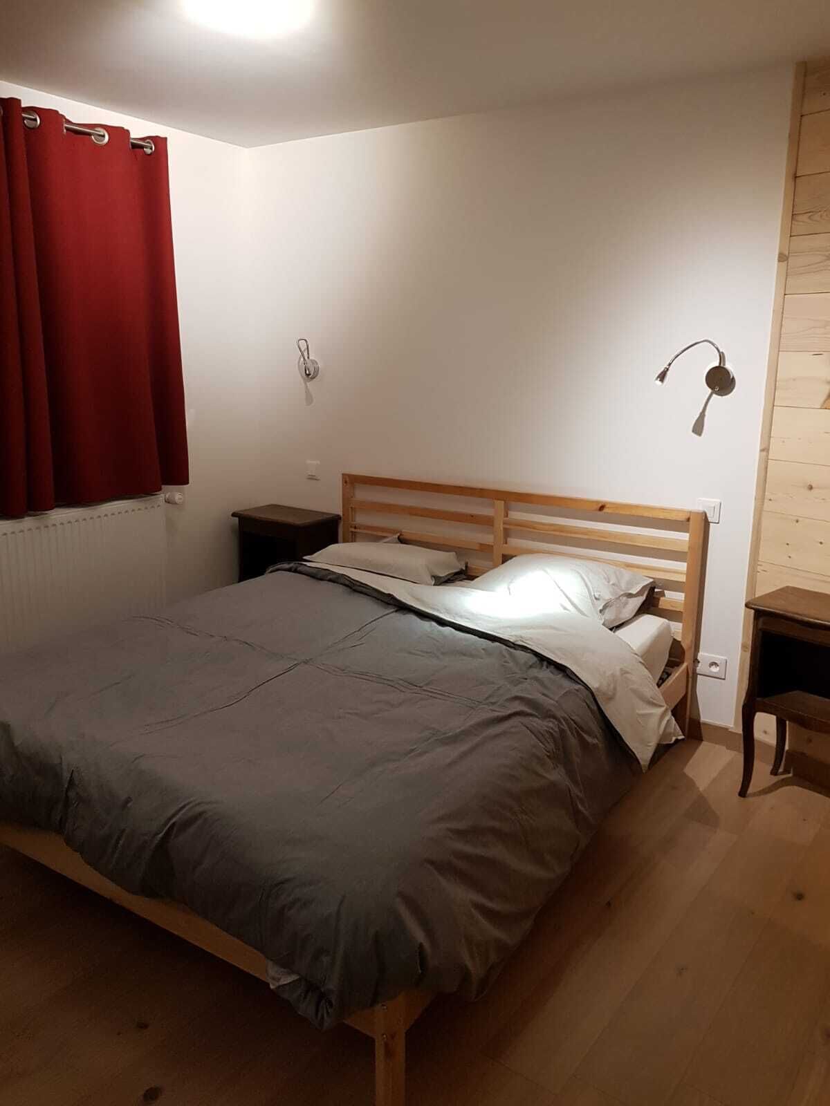 Un dormitori amb un llit de fusta, una tauleta de nit, una cortina vermella i un aplenç de llum fixat a la paret.