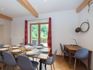 Un coin repas avec une table en bois dressée pour six personnes, entourée de six chaises. Une fenêtre avec des rideaux est visible et offre une vue sur la verdure à l’extérieur.