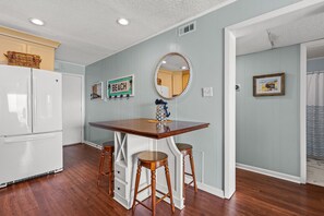 A kitchen with a wooden table, two stools, a refrigerator, and a wall-mounted mirror. - Amazing Panoramic Ocean, Marsh and Island Views in Secluded Vacation Condo Rental Resort (North Myrtle Beach)