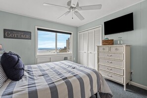A bedroom with a bed, a ceiling fan, a window with a view of the beach, a white dresser, and a flat-screen TV. - Amazing Panoramic Ocean, Marsh and Island Views in Secluded Vacation Condo Rental Resort (North Myrtle Beach)