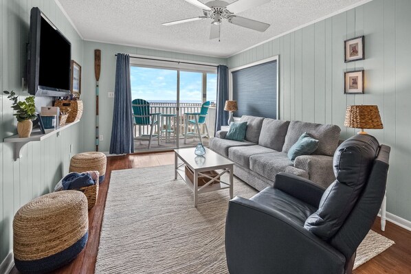A living room with a sofa, coffee table, and a TV mounted on the wall. There is a balcony with chairs and a view of the sky. - Amazing Panoramic Ocean, Marsh and Island Views in Secluded Vacation Condo Rental Resort (North Myrtle Beach)
