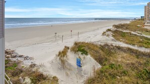 Una playa con un muelle a lo lejos, un cartel que indica acceso a la playa, y un edificio junto a la playa.