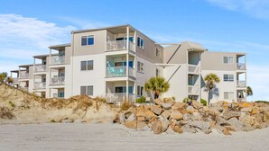 Un edificio frente al mar con varios pisos y balcones, una playa arenosa en primer plano y una barrera rocosa.