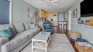 Una sala de estar con un sofá gris, un sillón azul y una mesa de centro blanca. Se observa una cocina al fondo con armarios de madera y un refrigerador.