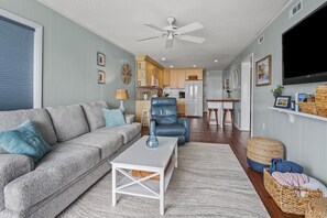 Ein Wohnzimmer mit grauem Sofa, blauem Sessel und weißem Couchtisch. Im Hintergrund ist eine Küche mit Holzschränken und einem Kühlschrank zu sehen.