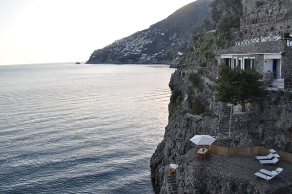 A cliffside hotel with a terrace overlooking the sea, featuring outdoor seating and a staircase leading to the building.