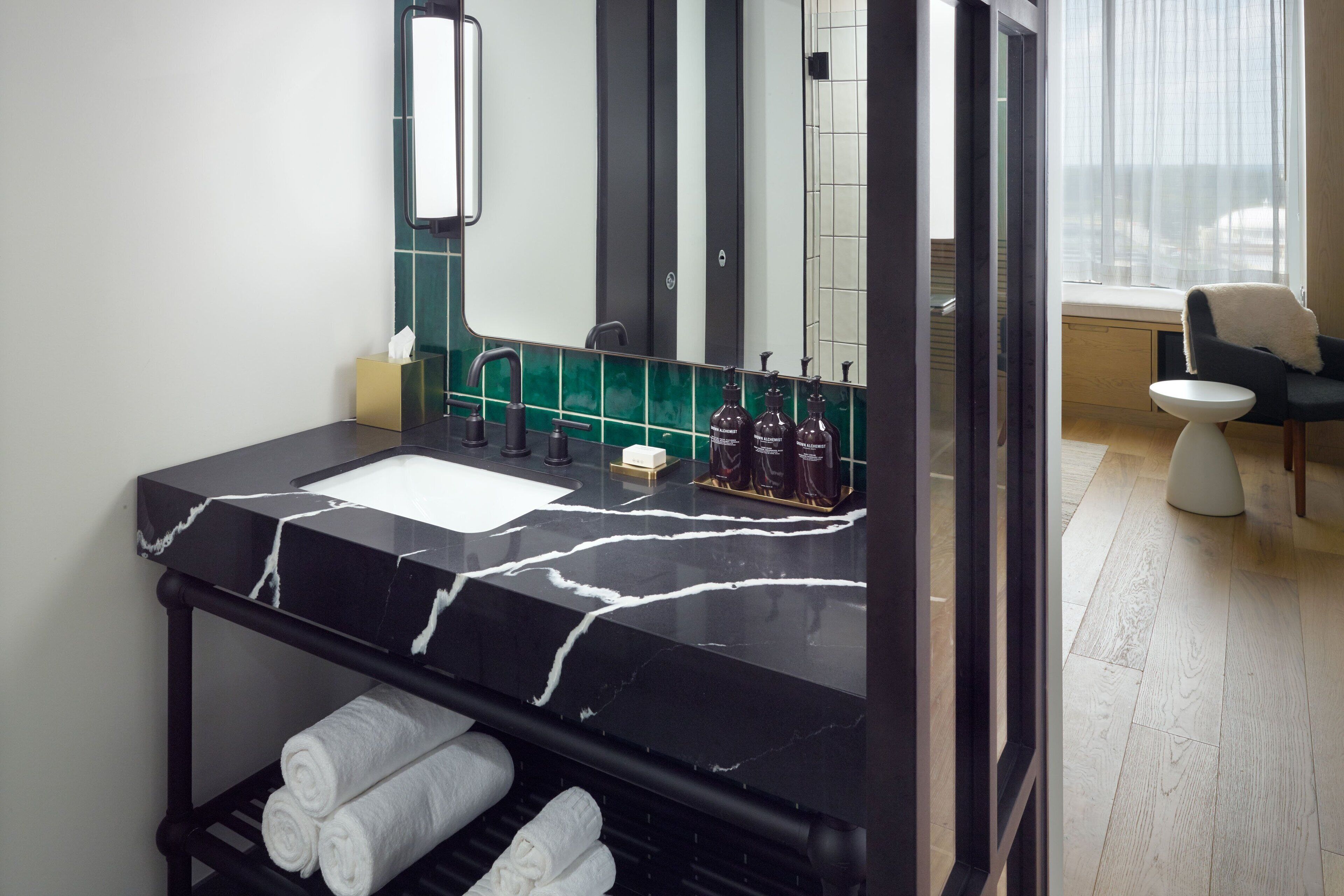 A modern bathroom with a black marble sink, green tiles, a large mirror, and a window with a view.