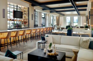 Zona de bar moderna con una barra larga, varios taburetes y una pantalla de televisión. Hay un área de estar con un sofá, sillas y una mesa de centro con adornos.