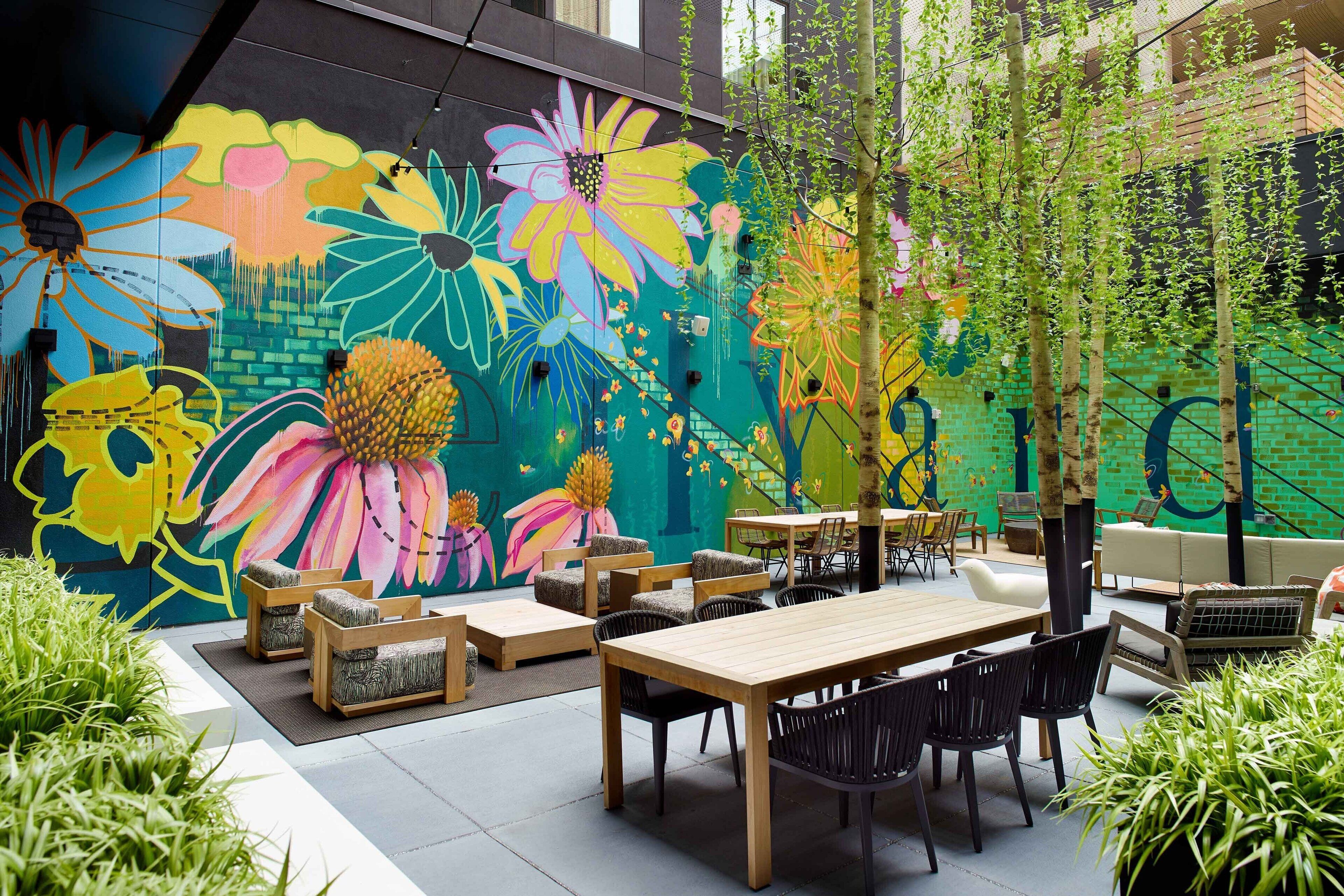 An outdoor seating area with a mural, wooden tables, and black chairs.