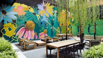 An outdoor seating area with a mural, wooden tables, and black chairs.