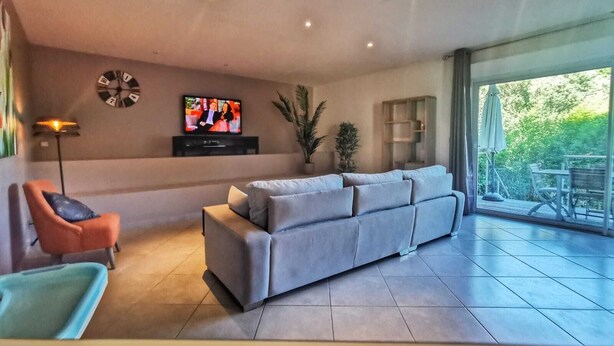 Een moderne woonkamer met een grijze bank, een televisie, een vloerlamp en uitzicht op het landschap door grote ramen.