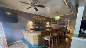 Een bar met een houten toonbank, barstoelen, een plafondventilator en een decoratieve hanglamp.