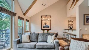 A cozy living room with a grey sofa, a brown leather chair, and a glass coffee table. There is a wooden ceiling with exposed beams, a chandelier, and a framed poster on the wall. - 2 Bedroom in Tarn Landing in Frisco (Frisco)