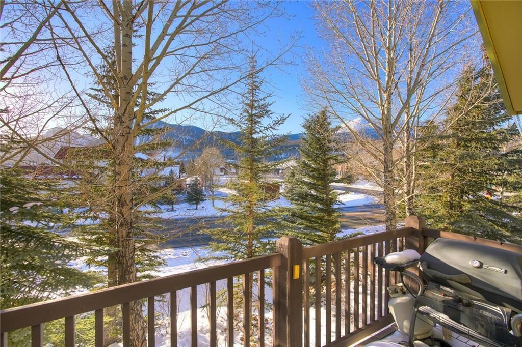 A deck with a barbecue grill, overlooking a snowy landscape with trees and distant mountains.