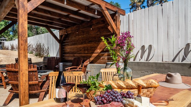 An outdoor dining area with wooden furniture, a table set with wine glasses, bread, grapes, and cheese.