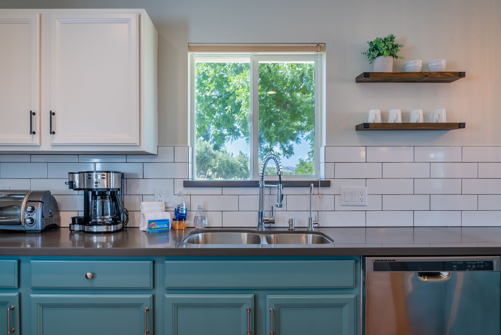 A modern kitchen with a coffee maker, toaster, and dishwasher. There is a window with a view of trees and a shelf with cups and a plant.